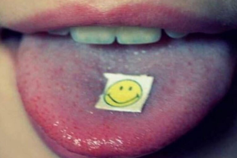Nahaufnahme einer Person, die einen Blotter mit einem Smiley-Gesicht auf der Zunge hält, was die Verwendung von legalen LSD-Analoga wie 1P-LSD, 1D-LSD, 1V-LSD und 1S-LSD symbolisiert.